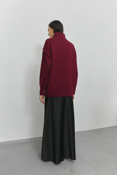 Femme portant un pull bordeaux à col roulé et une jupe longue noire, vue de dos, sur fond blanc minimaliste. Mode automne-hiver élégante.