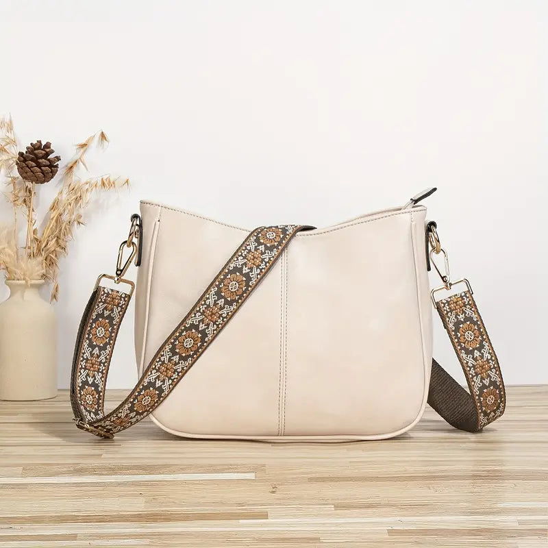 Sac à main en cuir beige avec bandoulière brodée florale, posé sur une table en bois clair, vase décoratif en arrière-plan. Accessoire mode élégant.