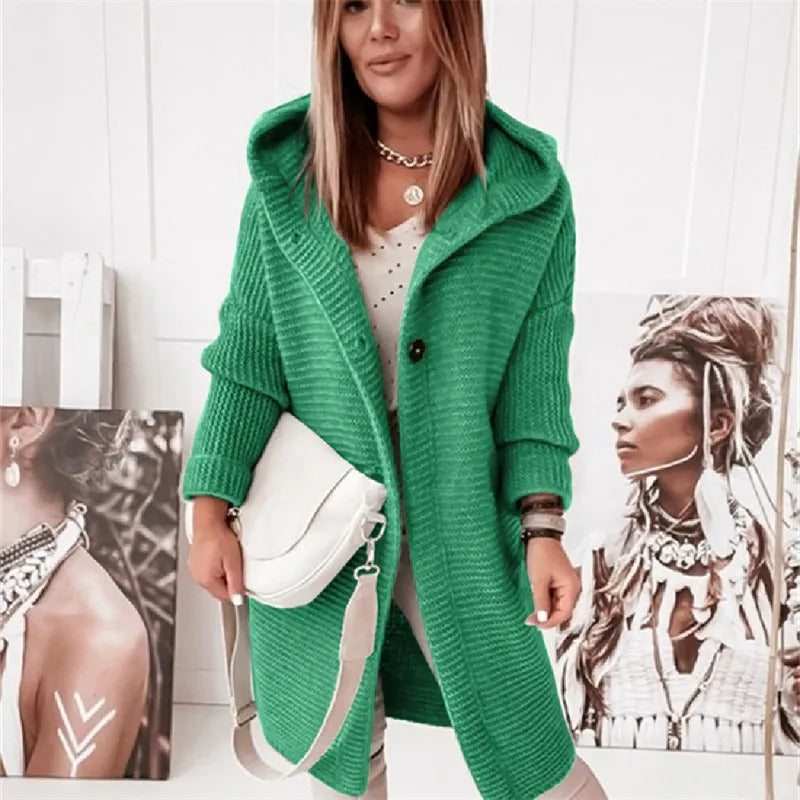 Femme portant un manteau vert en tricot avec capuche, posant devant des portraits artistiques. Mode automne, style décontracté, accessoires tendance.