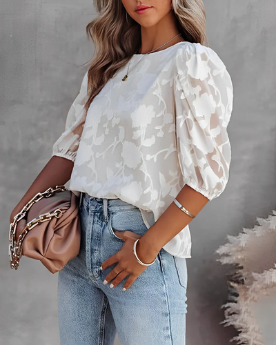 Femme portant un chemisier blanc à manches bouffantes et un jean, tenant un sac à main beige avec chaîne dorée. Mode élégante et tendance.