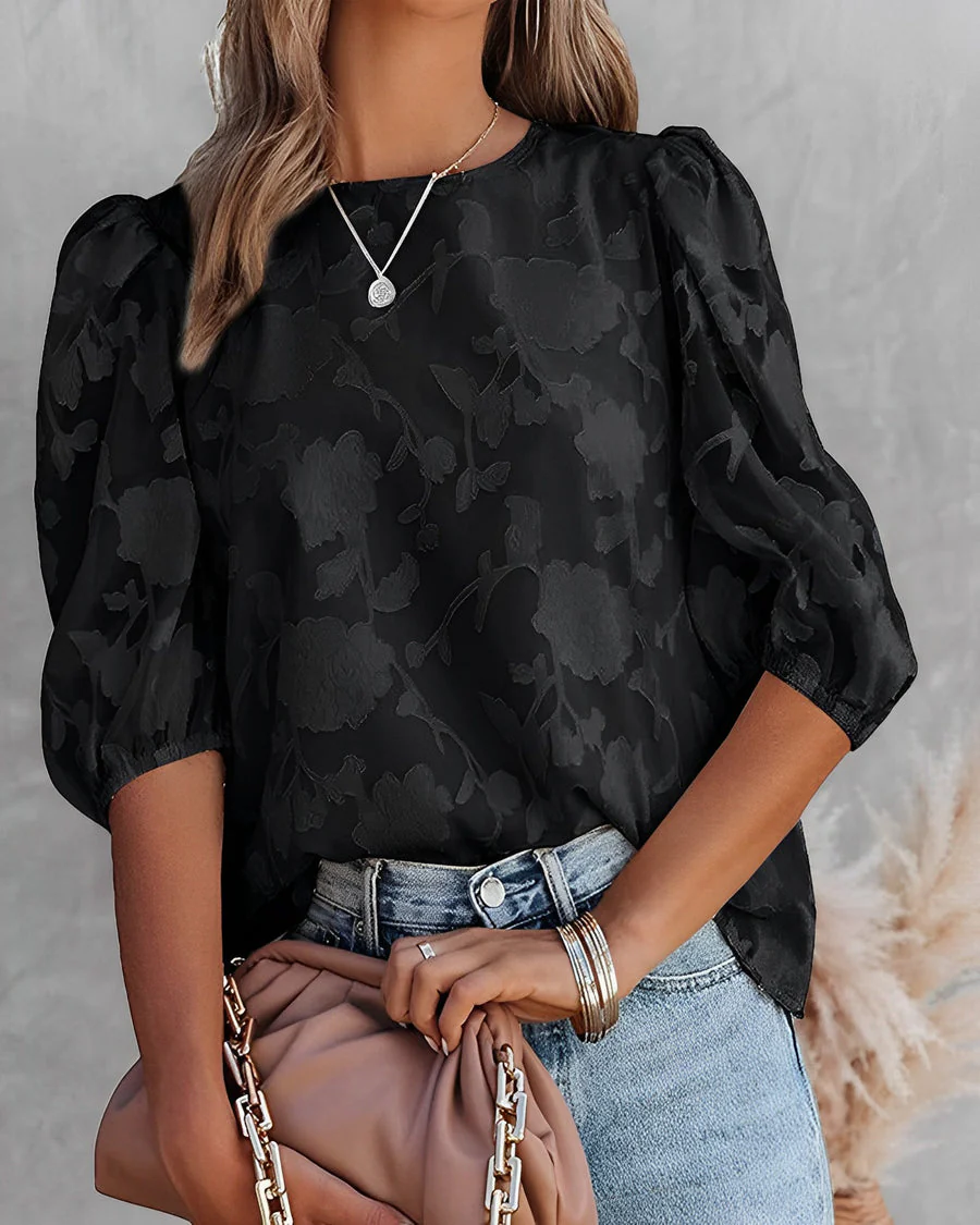 Femme portant un chemisier noir à motifs floraux, manches bouffantes, avec un collier argenté, tenant un sac beige. Mode élégante et tendance.