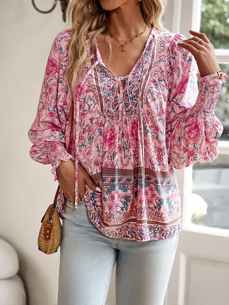 Femme portant un chemisier bohème rose à motifs floraux, manches longues, avec un sac en osier, debout près d'une fenêtre lumineuse.