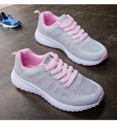 Chaussures de sport grises et roses pour femmes, semelle blanche, lacets roses, design respirant, posées sur un sol en bois.