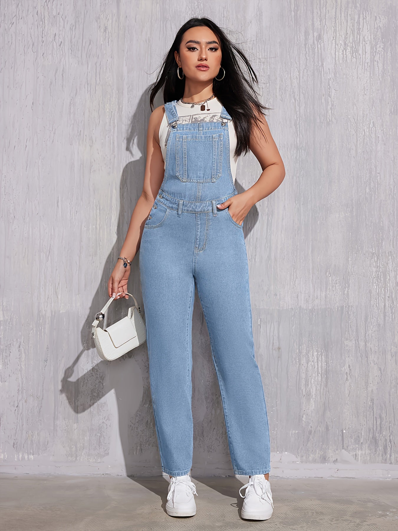 Femme portant une salopette en jean bleu clair, t-shirt blanc et baskets blanches, posant contre un mur gris avec un sac à main blanc.