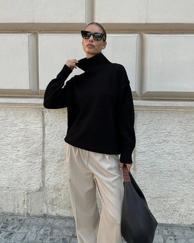 Femme élégante portant un pull noir à col roulé, pantalon beige ample, lunettes de soleil, sac à main noir, posant devant un mur texturé.