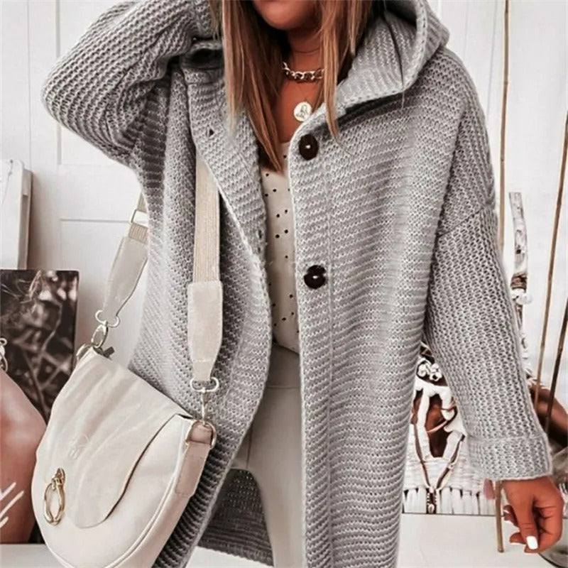 Femme portant un manteau en tricot gris avec capuche, boutons marron, et sac à main blanc. Mode automne-hiver, style décontracté chic.