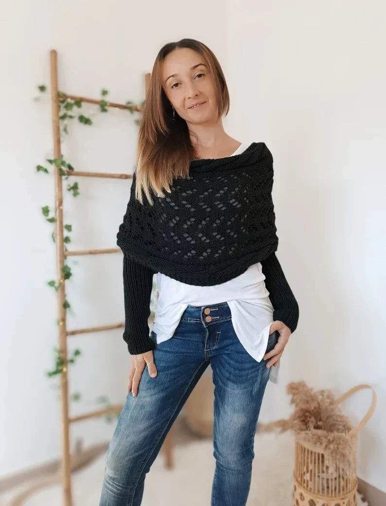 Femme portant un pull noir en tricot ajouré, debout devant une échelle en bois décorée de plantes vertes. Mode décontractée, style automne.