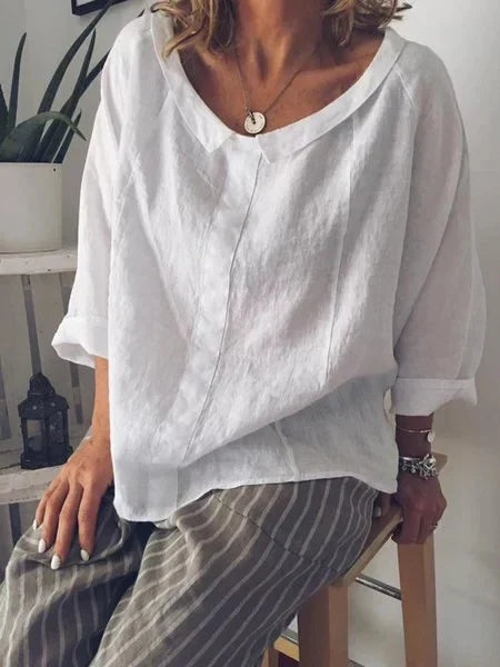 Femme portant une blouse blanche en lin, manches trois-quarts, assise sur un tabouret, avec un collier pendentif et un pantalon rayé.