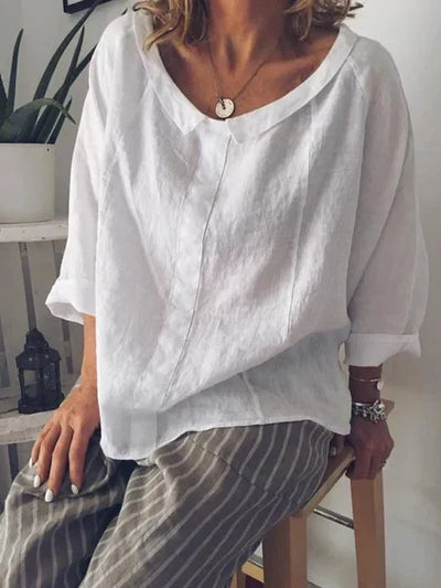 Femme portant une blouse blanche en lin, manches trois-quarts, assise sur un tabouret, avec un collier pendentif et un pantalon rayé.