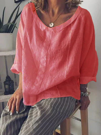 Femme portant un chemisier ample rose corail en lin, manches trois-quarts, assise sur un tabouret, avec un pantalon rayé gris et blanc.