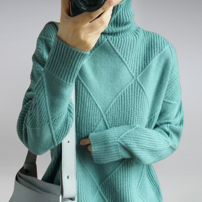 Pull en maille torsadée vert menthe avec col roulé, porté par une personne tenant un appareil photo. Sac à main gris en cuir visible. Mode automne-hiver.