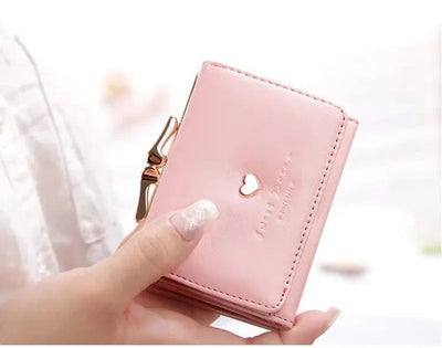 Portefeuille rose en cuir avec fermoir doré, motif cœur, tenu par une main féminine. Accessoire mode élégant et tendance pour femmes.