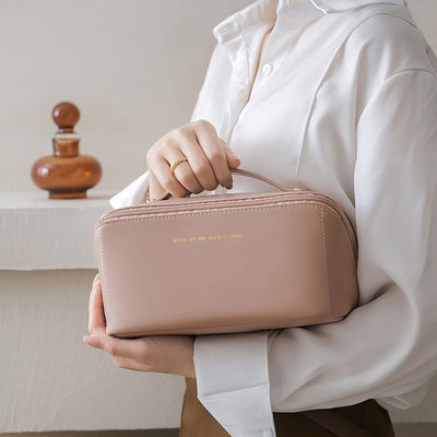 Femme tenant une élégante trousse de toilette rose en cuir, inscription dorée, sur fond de flacon décoratif. Accessoire de mode chic et pratique.