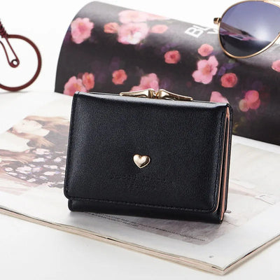Portefeuille noir en cuir avec motif cœur doré, posé sur un magazine avec lunettes de soleil. Accessoire élégant et tendance pour femmes.