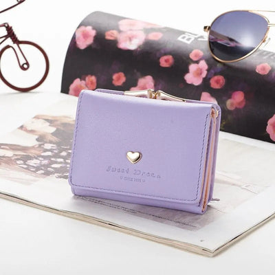 Portefeuille violet élégant avec cœur doré, posé sur un magazine avec lunettes de soleil et décoration florale en arrière-plan. Accessoire mode tendance.