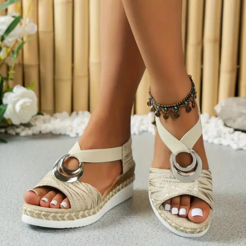 Sandales compensées beiges avec anneaux métalliques, portées par une femme avec un bracelet de cheville, sur fond de bambou et fleurs blanches.