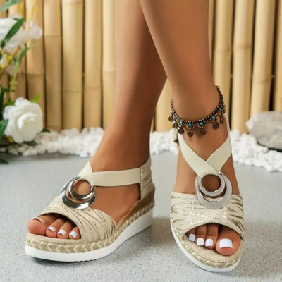 Sandales compensées beiges avec anneaux métalliques, portées par une femme avec un bracelet de cheville, sur fond de bambou et fleurs blanches.