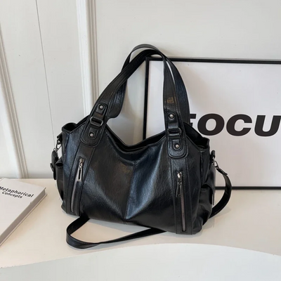 Sac à main en cuir noir élégant avec bandoulière, poches zippées, posé sur une table blanche. Accessoire mode femme chic et tendance.