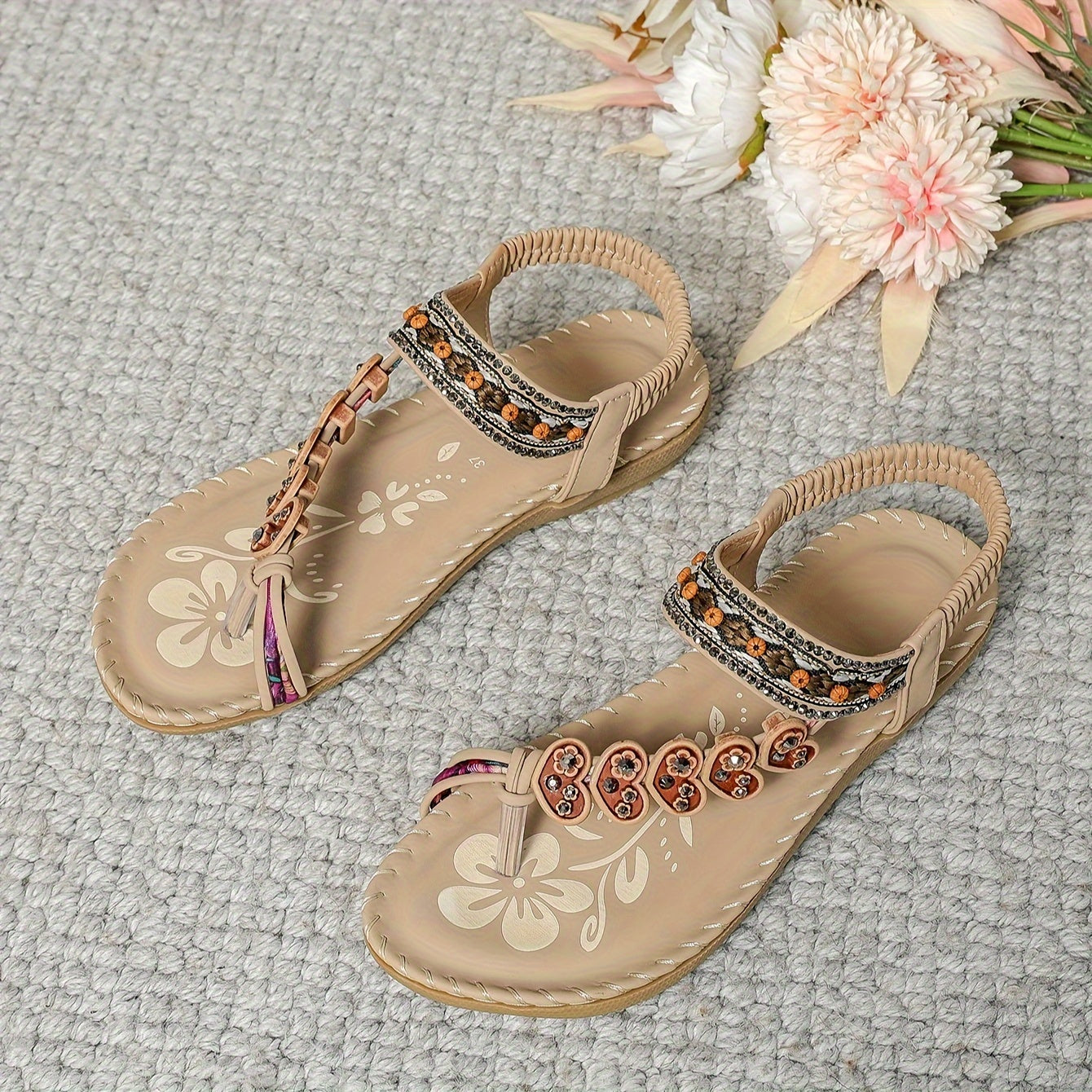Sandales d'été beiges pour femmes avec motifs floraux, ornées de perles orange et marron, posées sur un tapis gris, fleurs en arrière-plan.