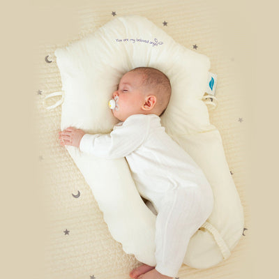 Bébé dormant paisiblement sur un coussin de soutien blanc, vêtement blanc, tétine verte. Literie étoilée, ambiance douce et sécurisée.