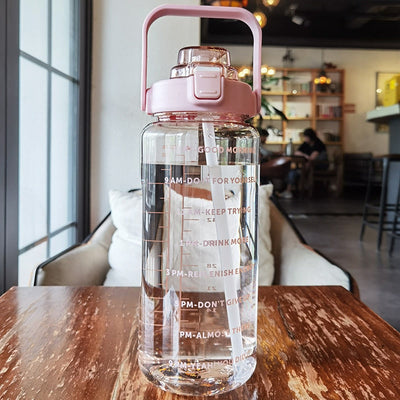 Bouteille d'eau transparente avec couvercle rose, graduations horaires et messages motivants, posée sur une table en bois dans un café moderne.