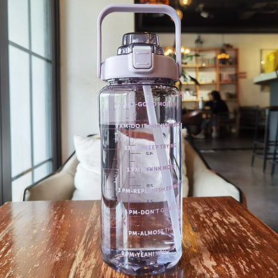 Gourde en plastique transparente avec paille intégrée, graduations horaires motivantes, posée sur une table en bois dans un café moderne.