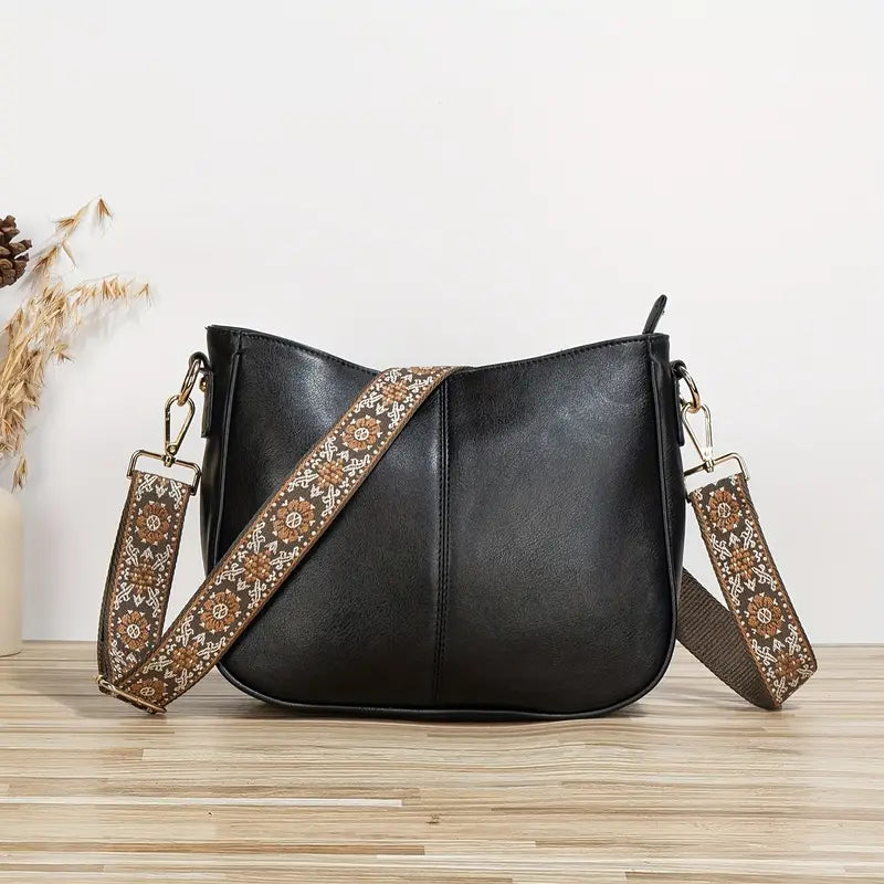 Sac à main noir en cuir avec bandoulière brodée florale marron, posé sur une table en bois clair. Accessoire mode élégant et tendance.