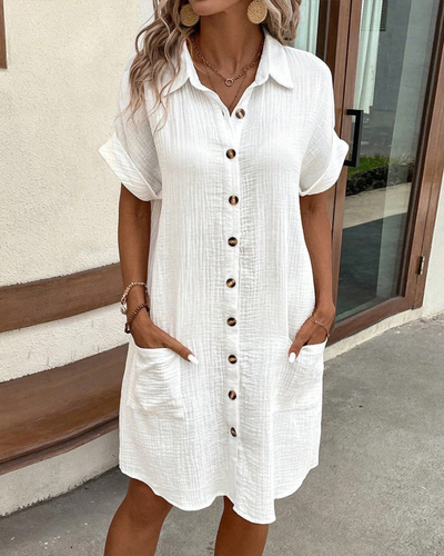 Robe chemise blanche en lin avec boutons en bois, manches courtes, poches latérales, portée par une femme devant une maison moderne.