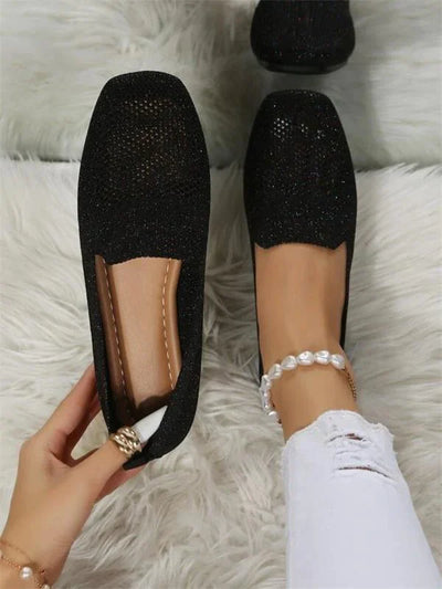 Chaussures plates noires élégantes pour femme, en tissu scintillant, portées avec un jean blanc déchiré et un bracelet de cheville perlé.