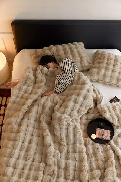Femme dormant sous une couverture en fausse fourrure beige sur un lit, avec coussins assortis et une table de chevet avec bougie allumée. Ambiance cosy.