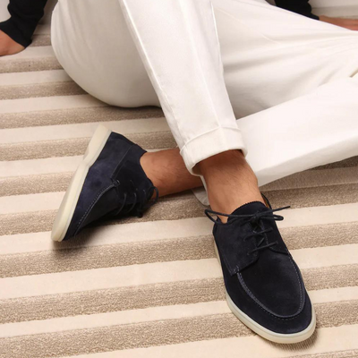 Chaussures bateau en daim bleu marine sur tapis rayé beige, portées avec un pantalon blanc. Style décontracté et élégant pour homme.