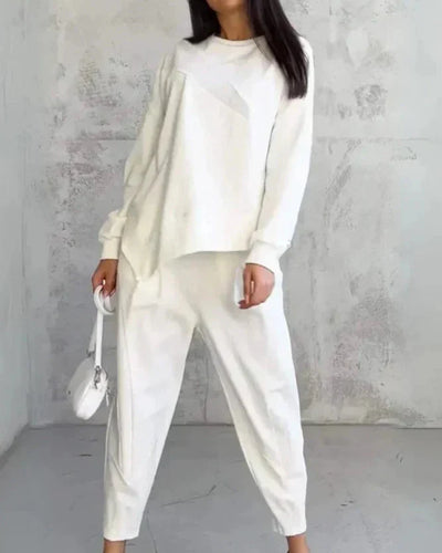 Femme en tenue décontractée blanche, portant un pull ample et un pantalon, avec un petit sac à main blanc, posant devant un mur texturé gris.