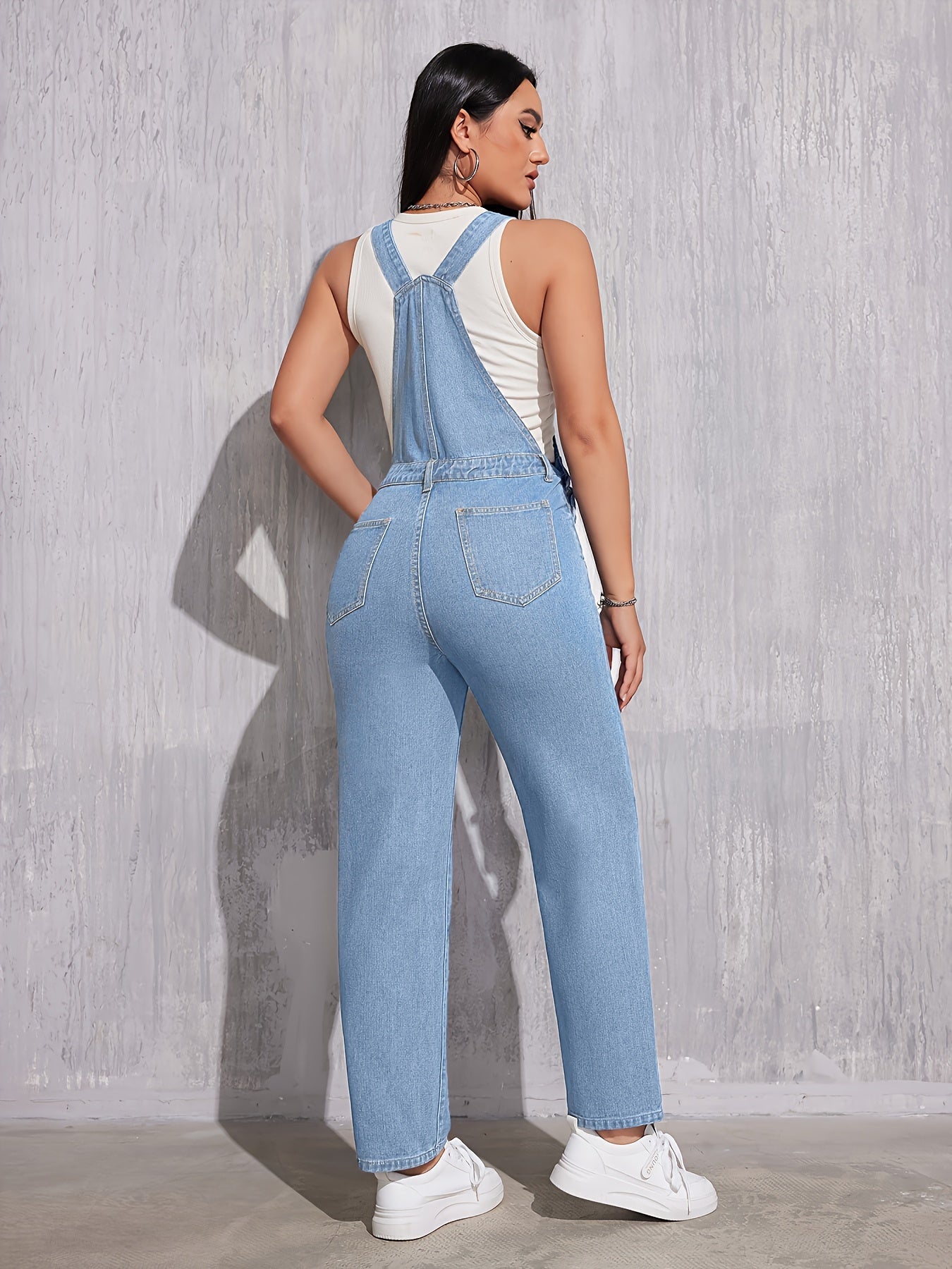 Femme portant une salopette en jean bleu clair, vue de dos, avec un débardeur blanc et des baskets blanches, posant devant un mur gris texturé.