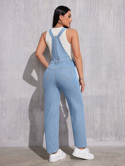 Femme portant une salopette en jean bleu clair, vue de dos, avec un débardeur blanc et des baskets blanches, posant devant un mur gris texturé.