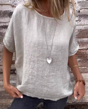 Femme portant un t-shirt en lin beige avec un collier pendentif cœur, posant devant un mur en brique. Mode décontractée, style estival.