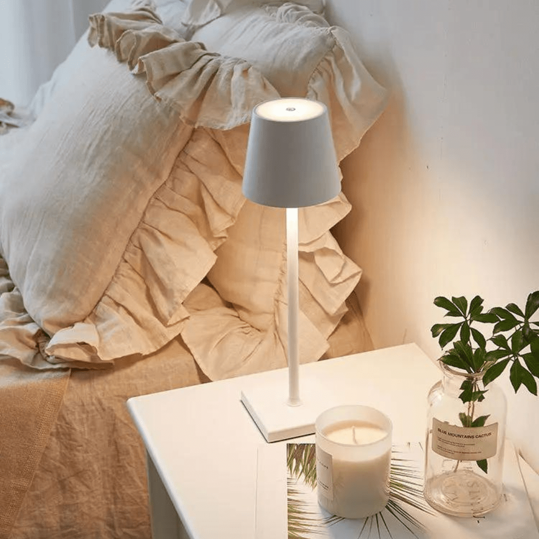 Lampe de chevet moderne blanche sur table de nuit, avec bougie parfumée et plante verte, à côté d'un lit avec coussins beiges en lin.