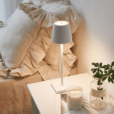 Lampe de chevet moderne blanche sur table de nuit, avec bougie parfumée et plante verte, à côté d'un lit avec coussins beiges en lin.