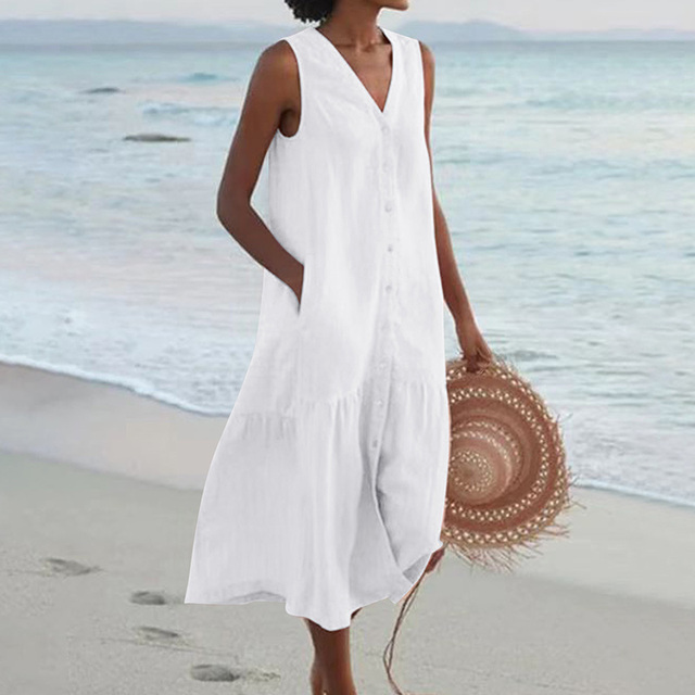 Robe blanche sans manches, femme tenant un chapeau de paille, marchant sur une plage au bord de la mer. Mode estivale, tenue décontractée plage.