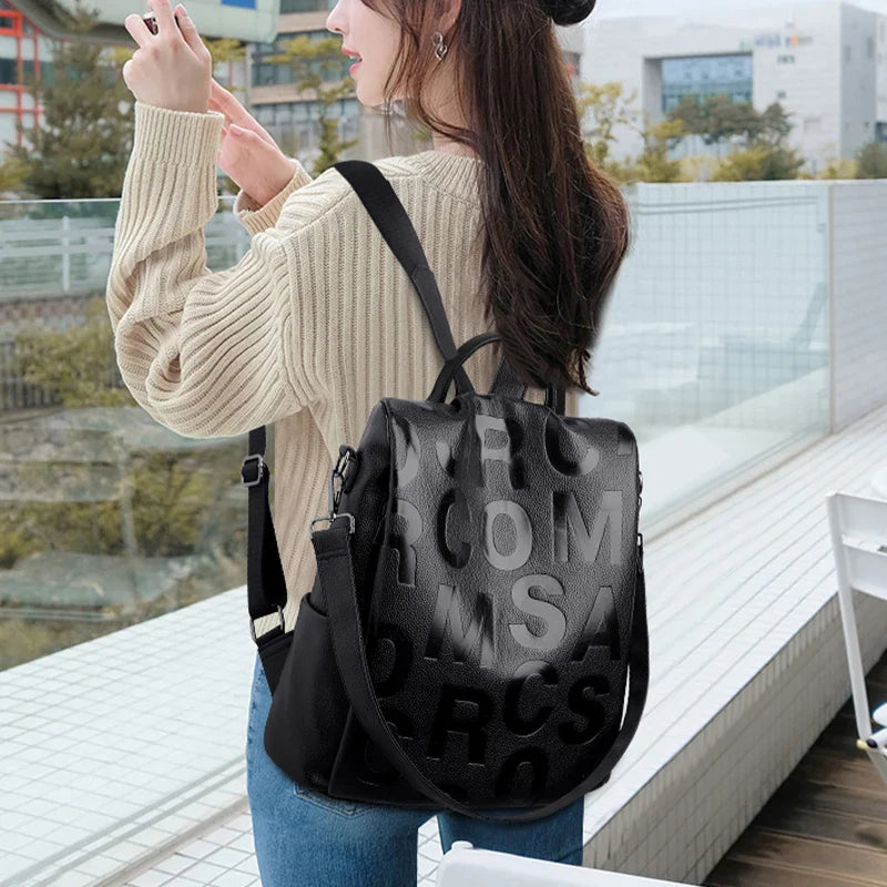 Jeune femme portant un sac à dos noir en cuir avec des lettres en relief, debout à l'extérieur. Mode urbaine, style décontracté, accessoires tendance.