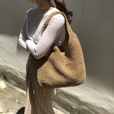 Femme portant un grand sac en paille tressée, tenue décontractée avec pull beige et jupe rayée, mode estivale chic et tendance.