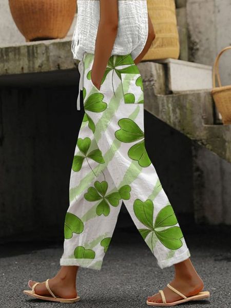Femme portant un pantalon blanc à motifs de trèfles verts, sandales beiges, haut blanc texturé, sac en osier en arrière-plan. Mode estivale décontractée.