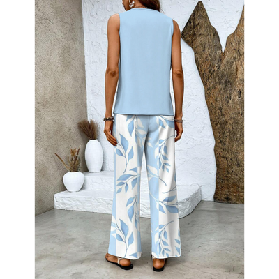 Femme en tenue estivale élégante, portant un haut bleu clair et un pantalon à motifs floraux bleus et blancs, debout près de pots décoratifs blancs.