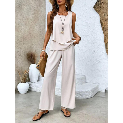 Femme en tenue d'été élégante, portant un ensemble blanc fluide, chapeau de paille, sandales, et sac à main, posant devant un mur texturé.