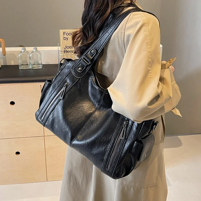 Femme portant un grand sac à main en cuir noir avec plusieurs poches zippées, vêtue d'un manteau beige, dans un intérieur moderne.