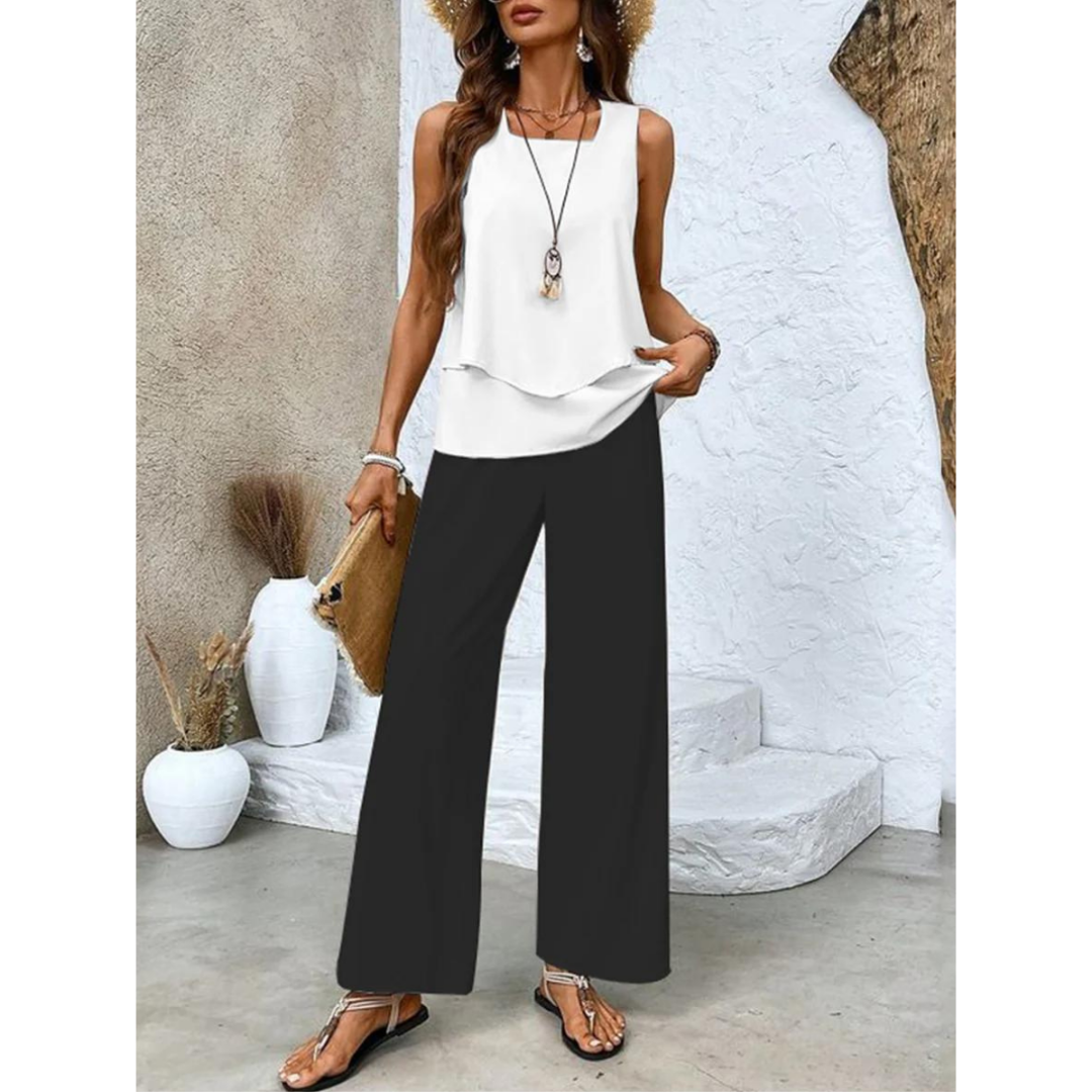 Femme en tenue estivale chic, portant un débardeur blanc, pantalon large noir, sandales, chapeau de paille, tenant un sac en paille, devant un mur texturé.