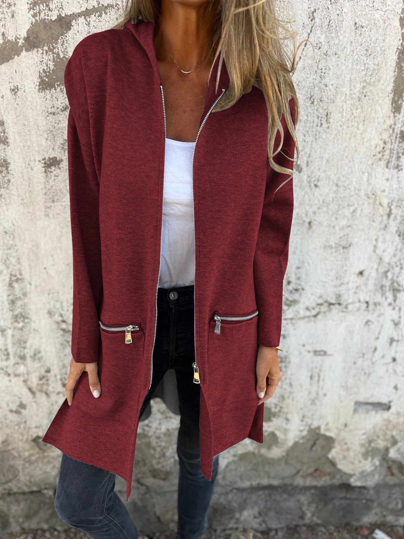 Femme portant un manteau rouge bordeaux avec fermetures éclair, sur fond de mur texturé. Mode automne, style décontracté, tendance vestimentaire.
