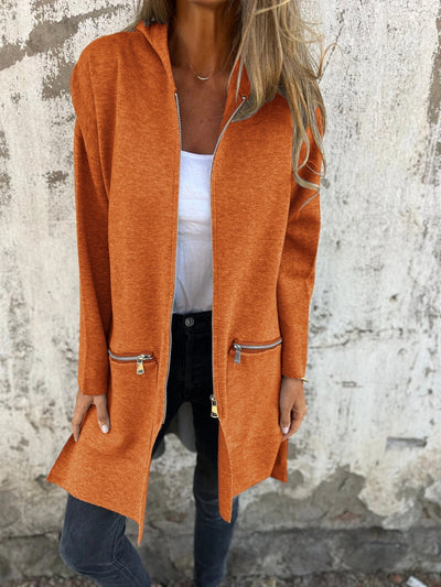 Femme portant un manteau orange à fermeture éclair, poches zippées, sur un mur texturé. Mode automne, style décontracté, tendance vestimentaire.