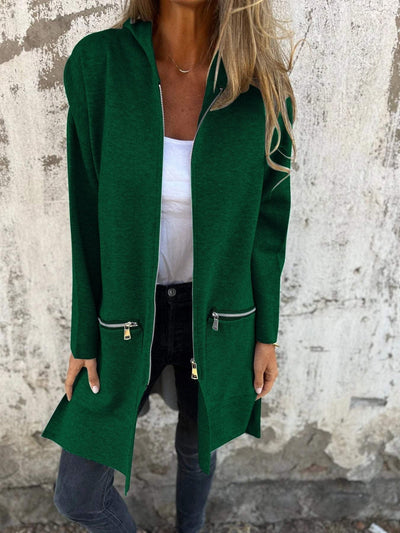 Femme portant un long cardigan vert avec poches zippées, t-shirt blanc et jeans noirs, posant devant un mur texturé. Mode automne chic.