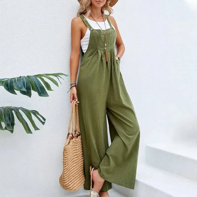 Femme en combinaison verte ample, portant un chapeau de paille et un sac en osier, posant contre un mur blanc. Mode estivale décontractée.