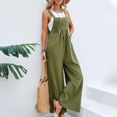 Femme en combinaison verte ample, portant un chapeau de paille et un sac en osier, posant contre un mur blanc. Mode estivale décontractée.